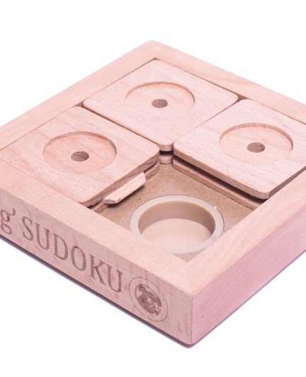 Sur la photo, un petit chien (ou un chat) est en train de jouer avec le PET' SUDOKU SMALL BASIC. Le jouet, un puzzle en bois avec plusieurs compartiments coulissants, est posé au sol. L'animal, concentré et curieux, utilise sa patte pour déplacer l'un des couvercles du puzzle dans le but de découvrir une friandise cachée à l'intérieur. Le fond de l'image est un intérieur chaleureux avec un sol en bois clair, accentuant la sensation de confort et de détente.